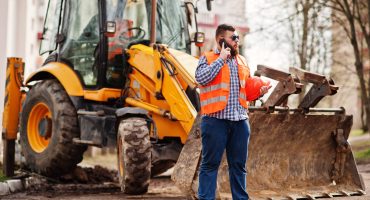 Beard worker man suit construction worker in safety orange helmet, sunglasses against tractor with mobile phone at hand.